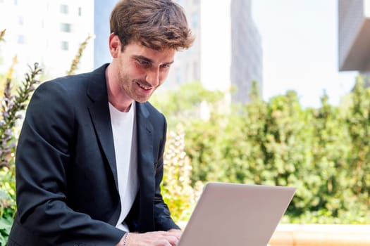 smiling young man using a laptop sitting on a bench of a park near of office buildings, concept of business and technology for remote work