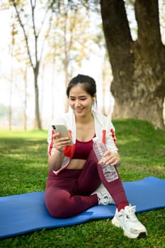 Cheerful woman relaxing after workout and using mobile phone, sitting on mat under trees in a sunny park.