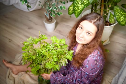 A young individual is comfortably seated on a soft surface, holding a vibrant plant surrounded by lush greenery in a bright room.