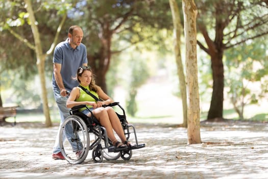 Nurse assisting a smiling young disabled woman in a wheelchair enjoying a sunny day at the park