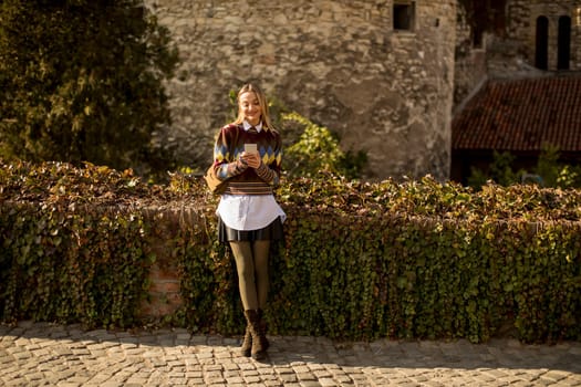Pretty young woman standing with mobile phone on street at sunny autumn day