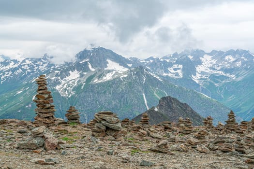 A breathtaking view of towering mountain peaks, adorned with patches of snow, contrasts with carefully stacked stone cairns on the rocky terrain. The clouds loom above, enhancing the atmosphere.