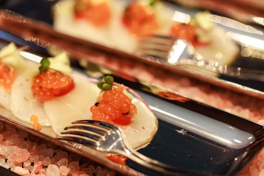 Elegant seafood appetizer featuring thinly sliced scallops, garnished with vibrant salmon roe, cherry tomatoes, microgreens, and a delicate sauce, artistically presented on sleek plates.