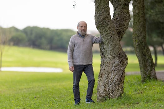 Portrait of a confident senior man relaxing in a green park, leaning against a cork oak tree, enjoying the peaceful atmosphere
