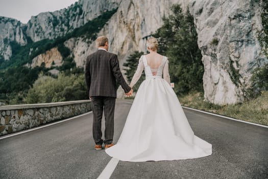 A bride and groom are standing on a road, holding hands. The bride is wearing a white dress and the groom is wearing a suit. The scene is set in a mountainous area, with a rocky road in the background