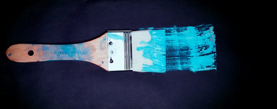 Large art brush with white bristles, paints a dark surface in bright turquoise, blue. Tool for artistic and restoration work, close-up view.