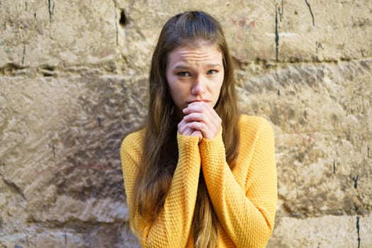 A young woman with long, flowing hair and a visibly anxious expression stands alone against a beautifully textured stone wall, clearly embodying deep feelings of fear, concern, and uncertainty
