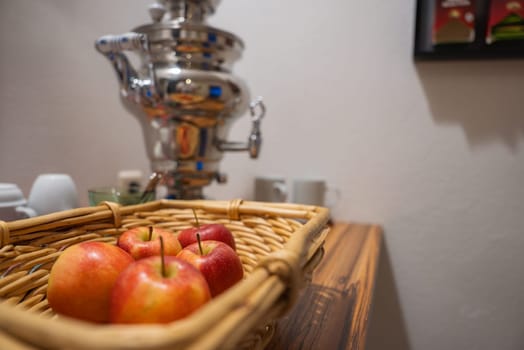 A wicker basket of red apples sits on a wooden surface with a silver samovar in the background, accompanied by white cups in a cozy, luxurious setting.