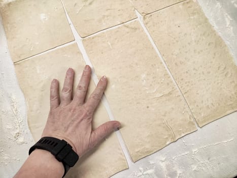 A hand is seen pressing down on evenly cut pieces of dough laid out on a floured countertop, indicating preparation for baking.