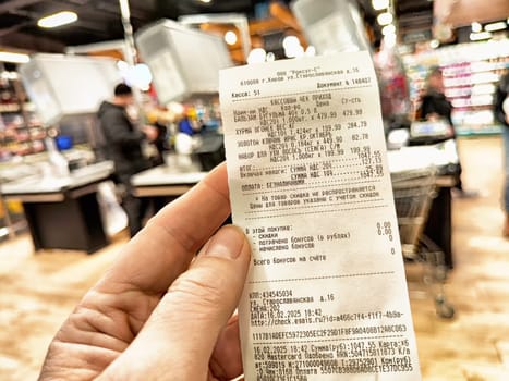 Kirov, Russia - February 14, 2025: A hand holds a detailed grocery receipt filled with various items bought at a bustling retail market during the day.