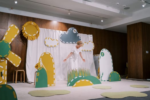 Actress stands on the stage of a kindergarten near the grass scenery, bending over with open arms. High quality photo