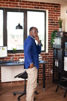 Black man with hand over mouth, standing in empty room and reflecting on startup business concepts. Male entrepreneur in brick wall workspace, contemplating ideas and observing office decor.