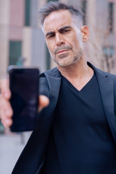 Confident Caucasian man in black outfit holding smartphone toward the camera with serious face. Business, technology, and communication concept in an urban environment with modern architecture.