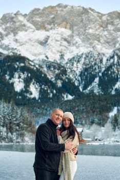 Romantic Winter Getaway Couple on Frozen Alpine Lake with Snowy Mountains and Rustic Cabin. Serene Outdoor Portrait of Man in Black Fleece and Woman in Cream Sweater Enjoying Picturesque Landscape - This high-resolution winter photograph captures a romantic couple enjoying a serene moment on a frozen alpine lake. The man, with short light brown hair, wears a black fleece jacket, dark pants, and white sneakers, while the woman, with long dark brown hair, is dressed in a beige knitted beanie, cream-colored chunky knit sweater, white leggings, and a long cream scarf. Their relaxed and content expressions convey a sense of peace and togetherness against the breathtaking winter backdrop. The background features snow-covered mountains densely forested with evergreen trees, their branches laden with snow. A rustic wooden cabin with a snow-covered roof nestles among the trees, adding a cozy element to the scene. The partially frozen lake reflects the surrounding landscape, creating a mirror-like effect that enhances the tranquil atmosphere. This image perfectly encapsulates the beauty of winter outdoor activities, romantic getaways, and the serene majesty of alpine landscapes, making it ideal for travel, lifestyle, and seasonal content.