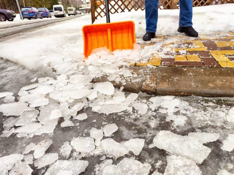 Snow and ice are cleared from the sidewalk using a shovel while cars remain parked along the street.