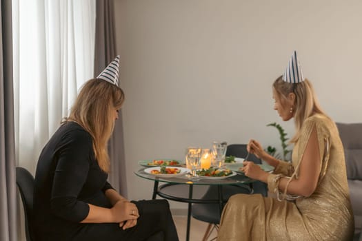 Two women, one wearing a party hat, sit at a small table with food and candles. One woman appears dejected the other eats. A quiet birthday dinner.