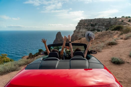 A red convertible car with two people in it, one of whom is waving. The car is driving on a road near the ocean