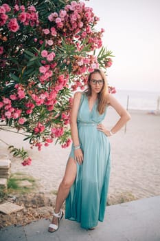 A graceful woman in a turquoise dress poses near blooming pink flowers, enjoying a peaceful moment in a natural outdoor setting.