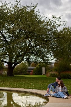 This picturesque scene captures two individuals sitting on the grass by the edge of a reflective pond in a serene park or garden. The couple is leaning against each other, enjoying a moment of relaxation surrounded by the vibrant colors of autumn. The background features trees adorned with red and orange leaves, and a building covered in lush green ivy. The reflection of the couple in the pond adds to the tranquil atmosphere, creating a beautiful and peaceful setting. Autumn Serenity: Couple Relaxing by a Reflective Pond in an Ivy-Covered Garden with Vibrant Foliage