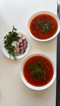 Traditional Ukrainian Borscht with Lard and Fresh Parsley. A top-down view of two bowls of traditional Ukrainian borscht served on a white table. The vibrant red beet soup is garnished with fresh parsley. Next to the bowls, a plate with sliced lard (salo) and fresh parsley adds to the authentic Eastern European meal. A classic homemade dish, rich in flavor and perfect for a hearty lunch or dinner.