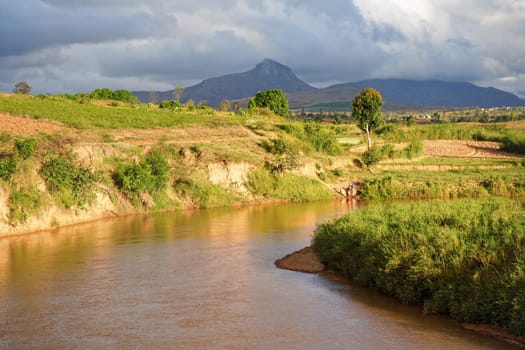 Sendrisoa, Madagascar - April 28, 2019: Slow muddy river, green grass and bushes on both sides, small mountains in background, woman washes clothes in water at left shore