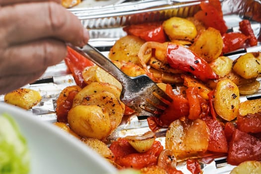Grilled potatoes and tomatoes served on aluminium foil tray straight from grill, closeup detail on silver fork in vegetables