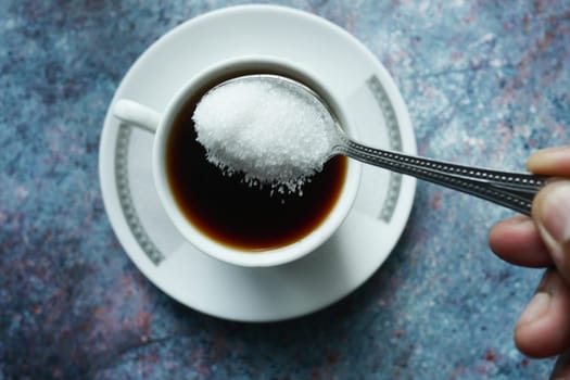 A hand holds a spoonful of sugar over a steaming cup of coffee.