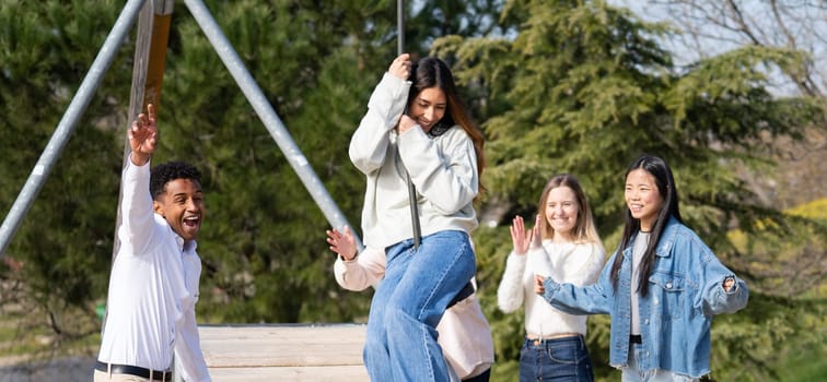 Group of cheerful young friends having fun in a park, one girl swinging on a zip line, the other two cheering her on