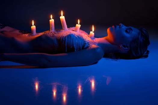 performance of tranquil naked female with burning candles and wax on slim body lying in studio with blue neon light