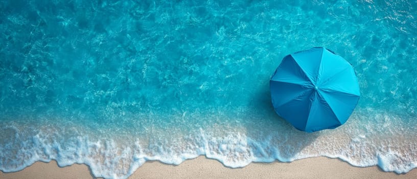 A blue beach umbrella stands near the shoreline, with ocean waves gently washing onto the sandy shore. A peaceful summer vacation scene.