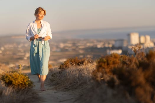 Woman enjoying a barefoot walk on a path overlooking a city at sunset, holding sunglasses and wearing a light dress