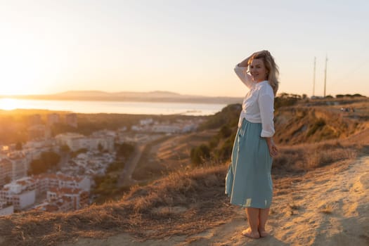 Blond woman standing barefoot on a hilltop, contemplating a beautiful cityscape by the sea at sunset