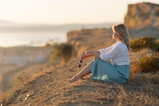 Blond woman sitting on a cliff at sunset holding sunglasses and enjoying the view
