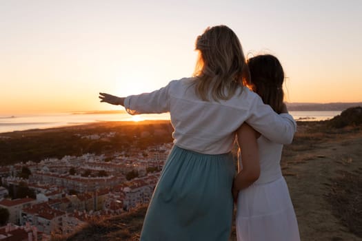 Mother and daughter sharing a tender moment, embracing as they gaze at the golden sunset over a picturesque coastal cityscape