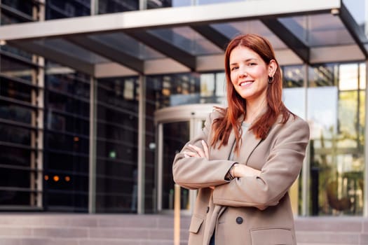 smiling young adult woman in casual suit standing looking at camera with arms crossed in front of an office building, concept of entrepreneur and business, copyspace for text