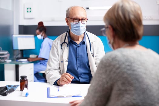 Senior woman and older male physician seated at desk, discussing diagnosis results from clipboard during checkup. Retired doctor in face mask is providing medical advice to female patient in clinic.
