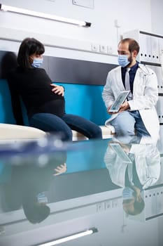 Doctor shares maternity advice with pregnant woman seated beside him, reviewing medical results on digital device. Male physician explains prenatal care to expectant lady, using tablet for visual aid.