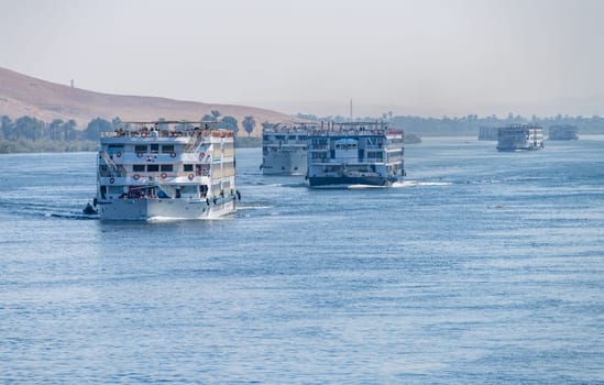 Esna, Egypt - 3 November 2024: Multiple river cruise ships make their way down the Nile towards Esna with desert in background