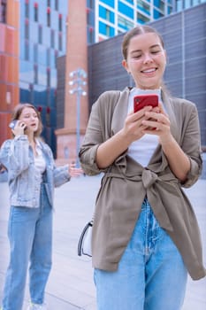 Two young caucasian women using smartphones outside. One texting and one talking on the phone, casual outfits, tall buildings in background. Connected, digital and urban lifestyle scene. Vertical