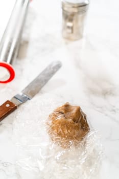 Front angled close-up view of a ball of amber gingerbread dough wrapped in plastic wrap, resting on a floured marble surface to chill before rolling.
