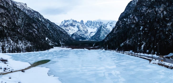A frozen lake in the Alps with clear icy patches revealing greenish water, surrounded by snow covered peaks, dense forests, and a narrow road.