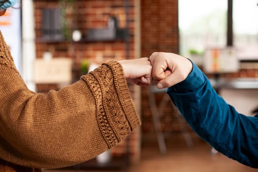 Business partners in startup office celebrate their collaboration with fist bump. Selective focus on hands of caucasian coworkers safely greeting each other in brick wall room.