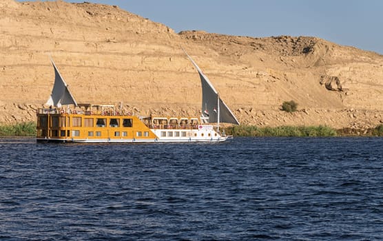 Edfu, Egypt - 3 November 2024: Small river cruise boat Amunet sails down the Nile with Sahara desert behind