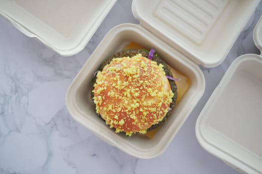 A vibrant burger topped with yellow flakes sits in a takeaway box.