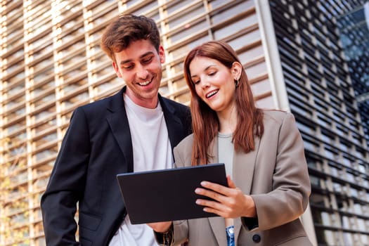 man and a woman smiling happy looking digital tablet together outdoors in front of an office building, the man is wearing a suit and the woman is wearing a jacket, concept of business and technology