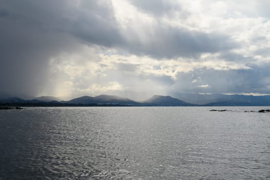 A breathtaking view unfolds as dark clouds loom over a serene body of water. Rays of sunlight break through, illuminating the distant mountains. A moment of nature's splendor captured during midday.