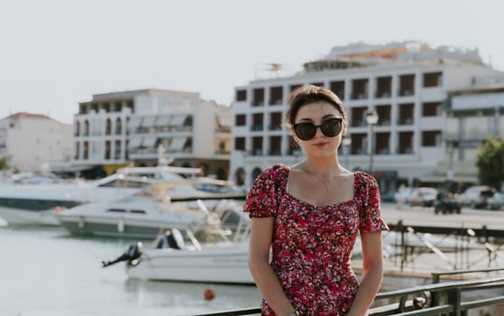 A portrait of one young beautiful Caucasian brunette girl in sunglasses and a bright colorful dress stands on the right, leaning against an iron railing against the backdrop of a blurred embankment with boats on a sunny summer day and looks smiling at the camera, close-up side view.