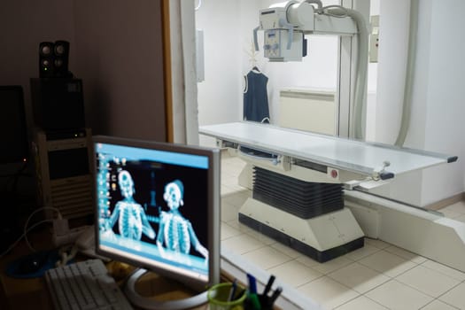 A medical X-ray room viewed from the control station, featuring an X-ray machine, a patient table, and a monitoring computer.The setup includes a protective glass window, a control panel, and office supplies on the desk.