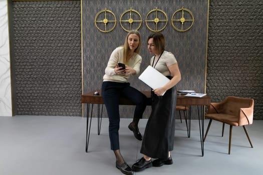 Two women engage in a collaborative discussion in a contemporary workspace. One shows content on a smartphone while the other leans on a table holding a tablet, surrounded by a chic backdrop.