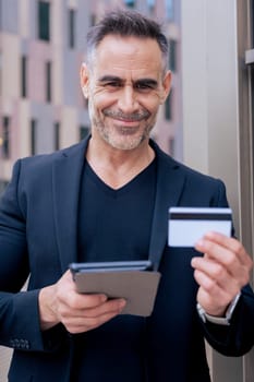 Confident caucasian man with headphones smiling while using tablet and holding a credit card outdoors. Concept of online shopping, digital payment, modern lifestyle, and financial technology. Vertical